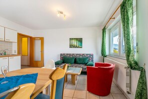 Een kleine kamer met een eettafel, stoelen, een bank, een rode stoel, een radiator en een raam met gordijnen.