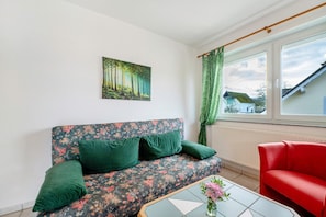 Een kamer met een bloemenbank, een rode fauteuil, een boslandschap op een schilderij, een raam met gordijnen en een betegelde salontafel.
