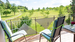 Ein Balkon mit zwei Stühlen und einem Tisch, der auf eine grüne Landschaft mit Bäumen und einem Weg blickt.
