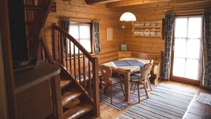 L’intérieur d’un chalet accueillant, avec un escalier en bois, une table à manger entourée de chaises, un lustre et une fenêtre agrémentée de rideaux.