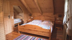 Une chambre dans une cabane en bois, avec un lit, deux tables de chevet, une petite table, une fenêtre avec des rideaux et un plafond en bois.