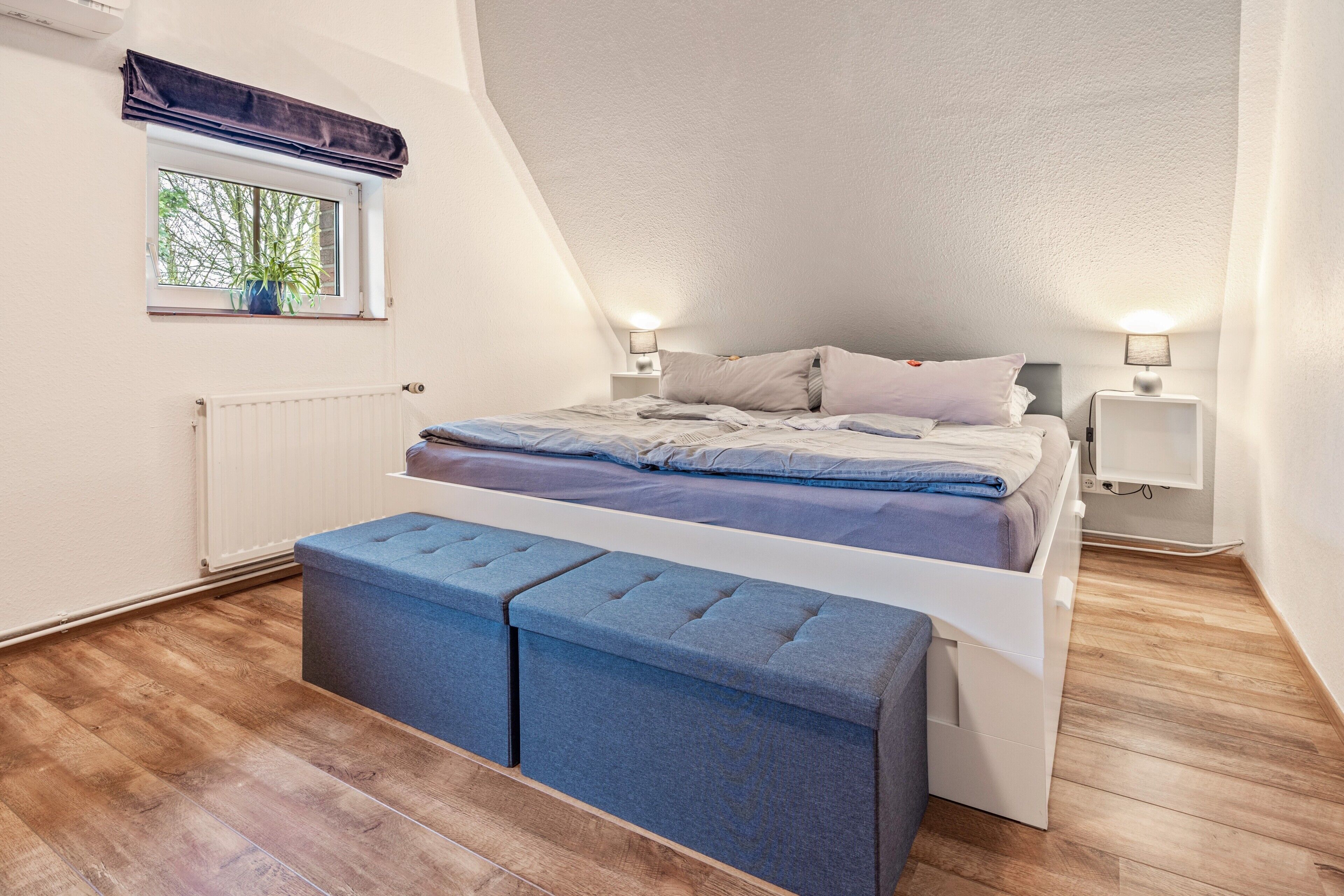 Un dormitorio con techo inclinado, una ventana con planta, una cama con almohadas, dos mesitas de noche con lámparas y un banco al pie de la cama.