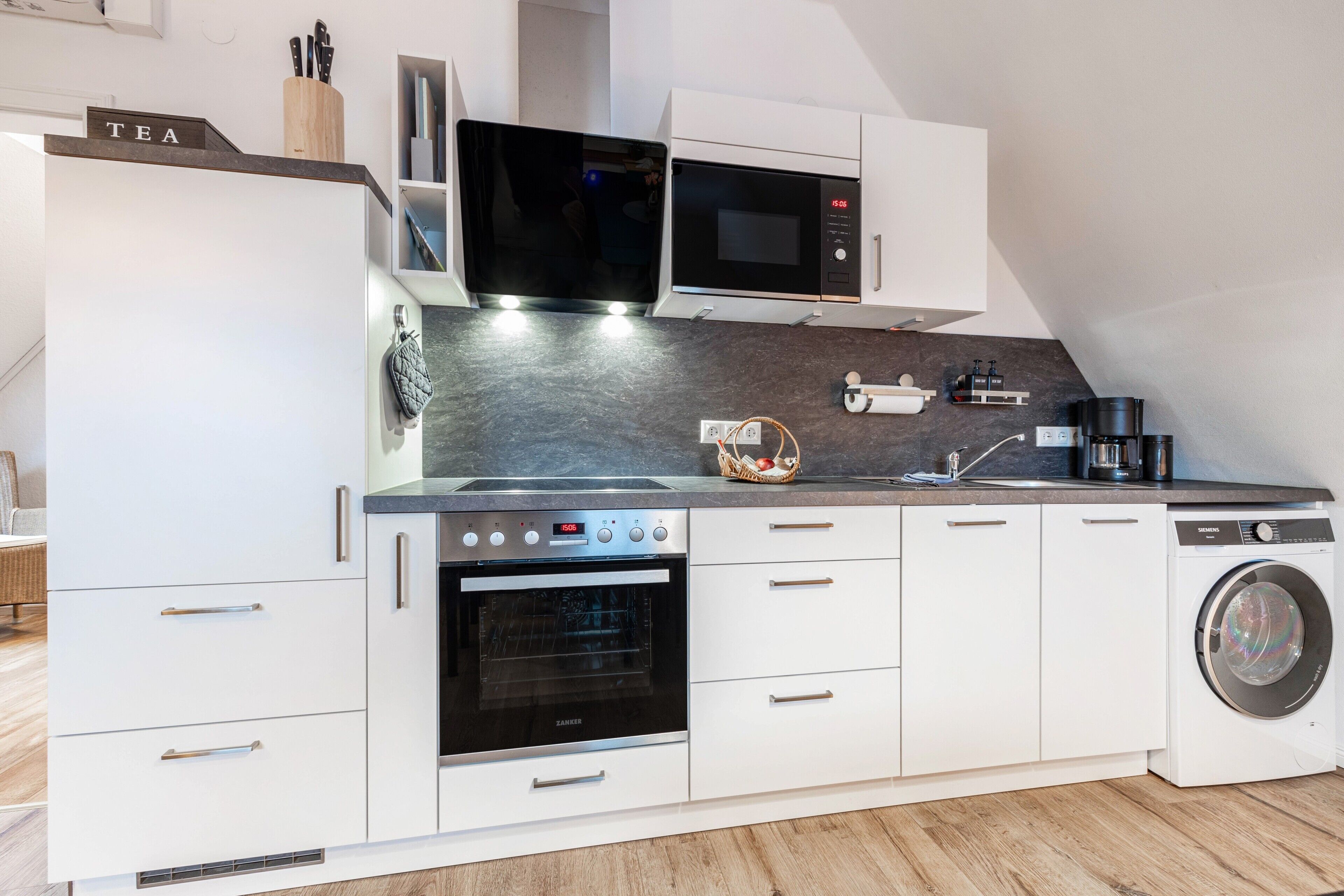 A modern kitchen with white cabinetry, a built-in oven, a microwave, a coffee maker, and a washing machine.