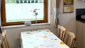 Een eethoek met een tafel gedekt voor twee personen, een raam met uitzicht op groen, een klok en een prikbord.