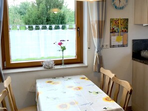 A dining area with a table set for two, a window with a view of greenery, a clock, and a bulletin board.