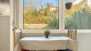 Mesa com toalha branca, cadeiras de madeira, planta em vaso e vista para uma parede de pedra e vegetação pela janela.