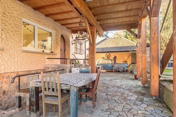 Une terrasse extérieure couverte, avec une table et des chaises en pierre, un banc en bois, et une voiture garée à l’arrière-plan.