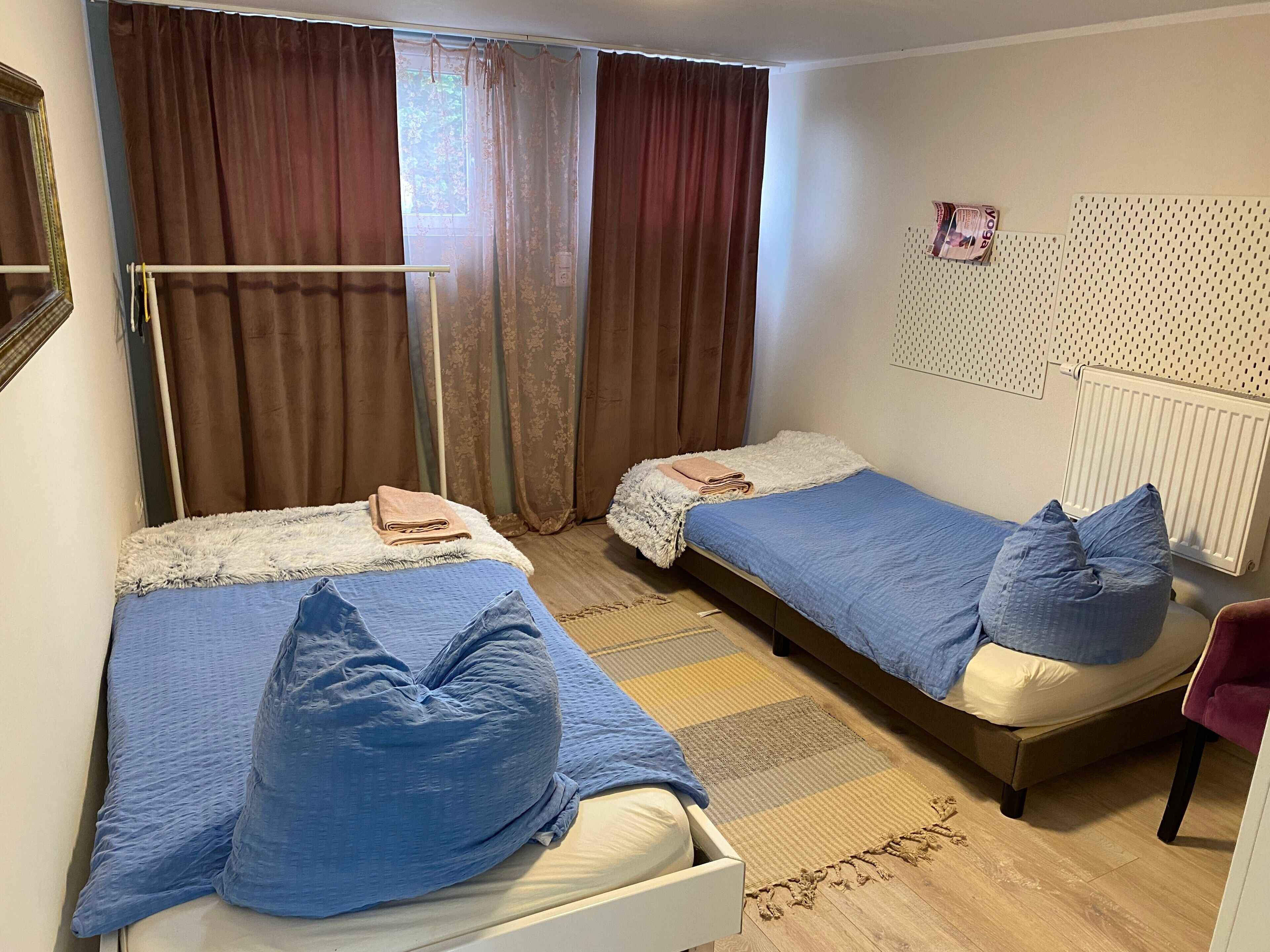 Una habitación con dos camas, una ventana con cortinas, un radiador y una lámpara fijada a la pared.