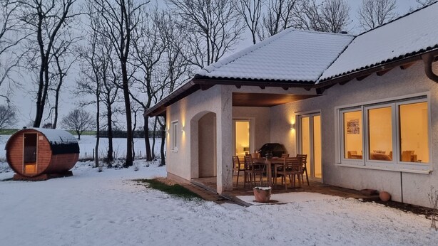 Uma cabana acolhedora com fachada de madeira, área de jantar com cadeiras e um grande barril na neve.