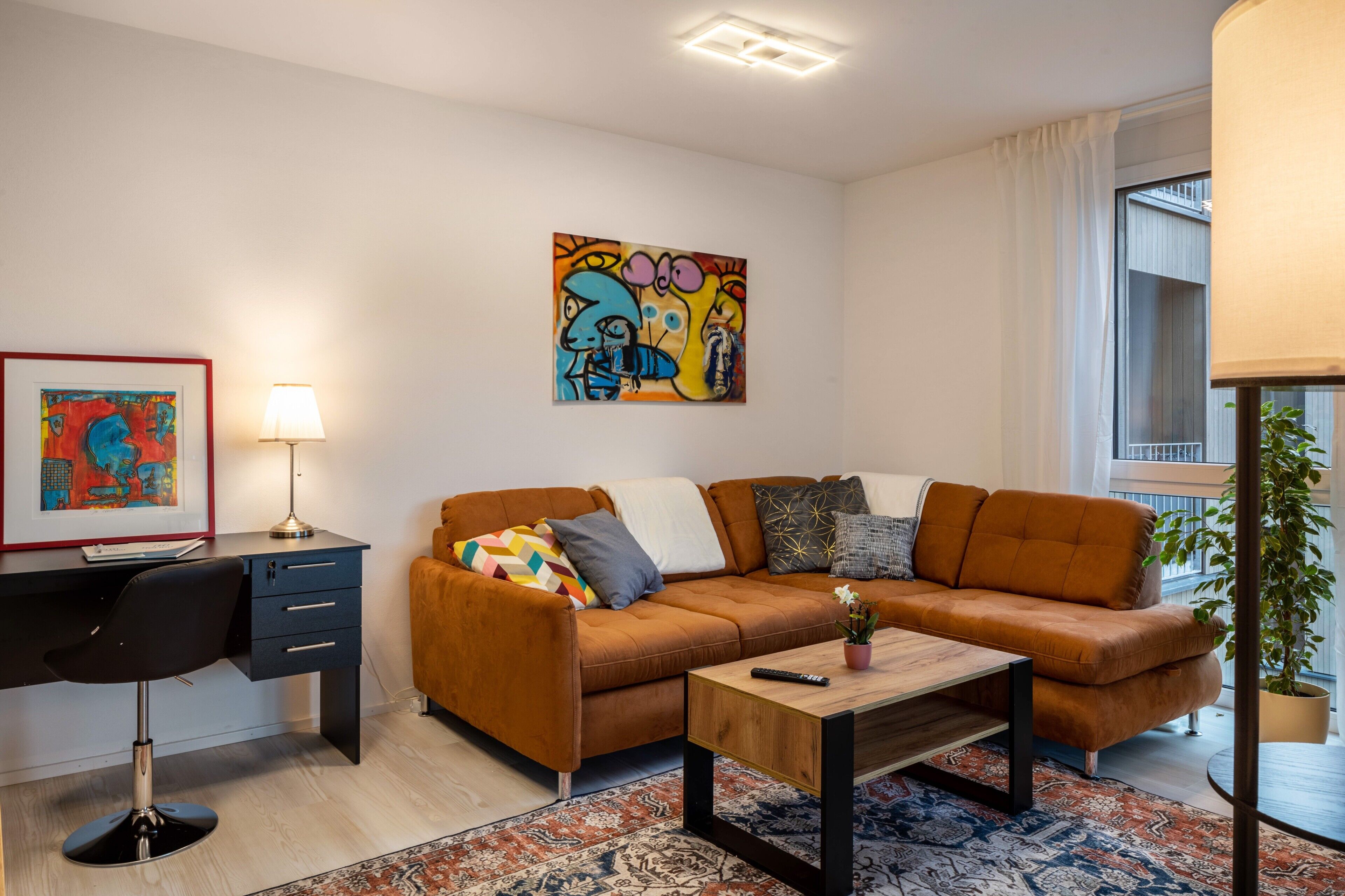 Un salon moderne comprenant un canapé, un bureau, une chaise, une table basse, un tableau et une fenêtre avec une plante.
