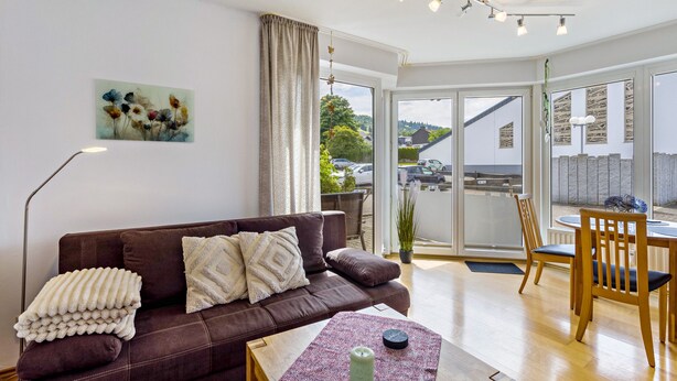 A living room with a brown sofa, a coffee table, a dining table with chairs, a large window, and a painting on the wall.