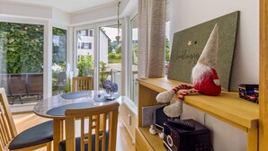 A dining area with a round table and chairs, a large window with a view of greenery, a shelf with a plush toy, and a framed picture on the wall.