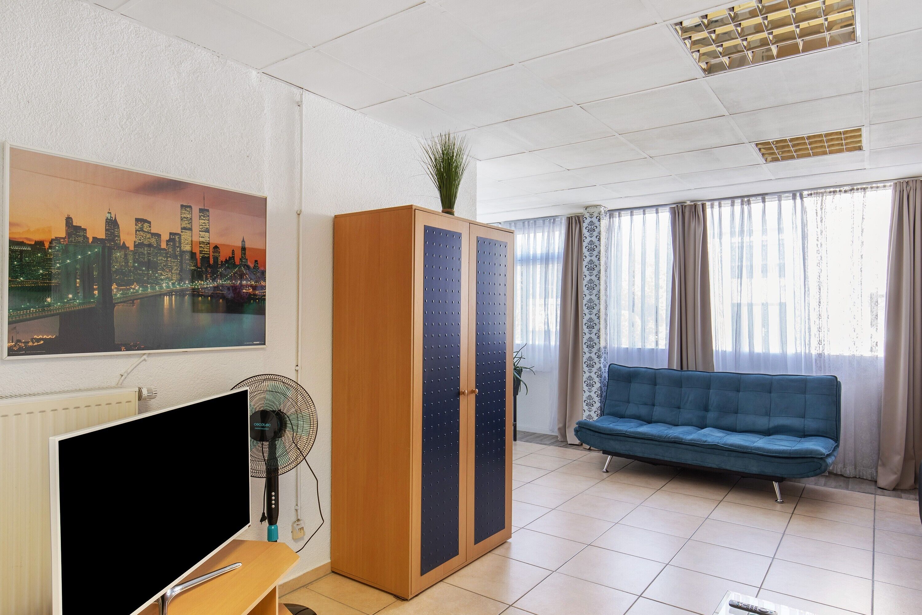 Een kamer met een televisie, een houten kledingkast, een blauwe bank en een stadsgezicht schilderij.
