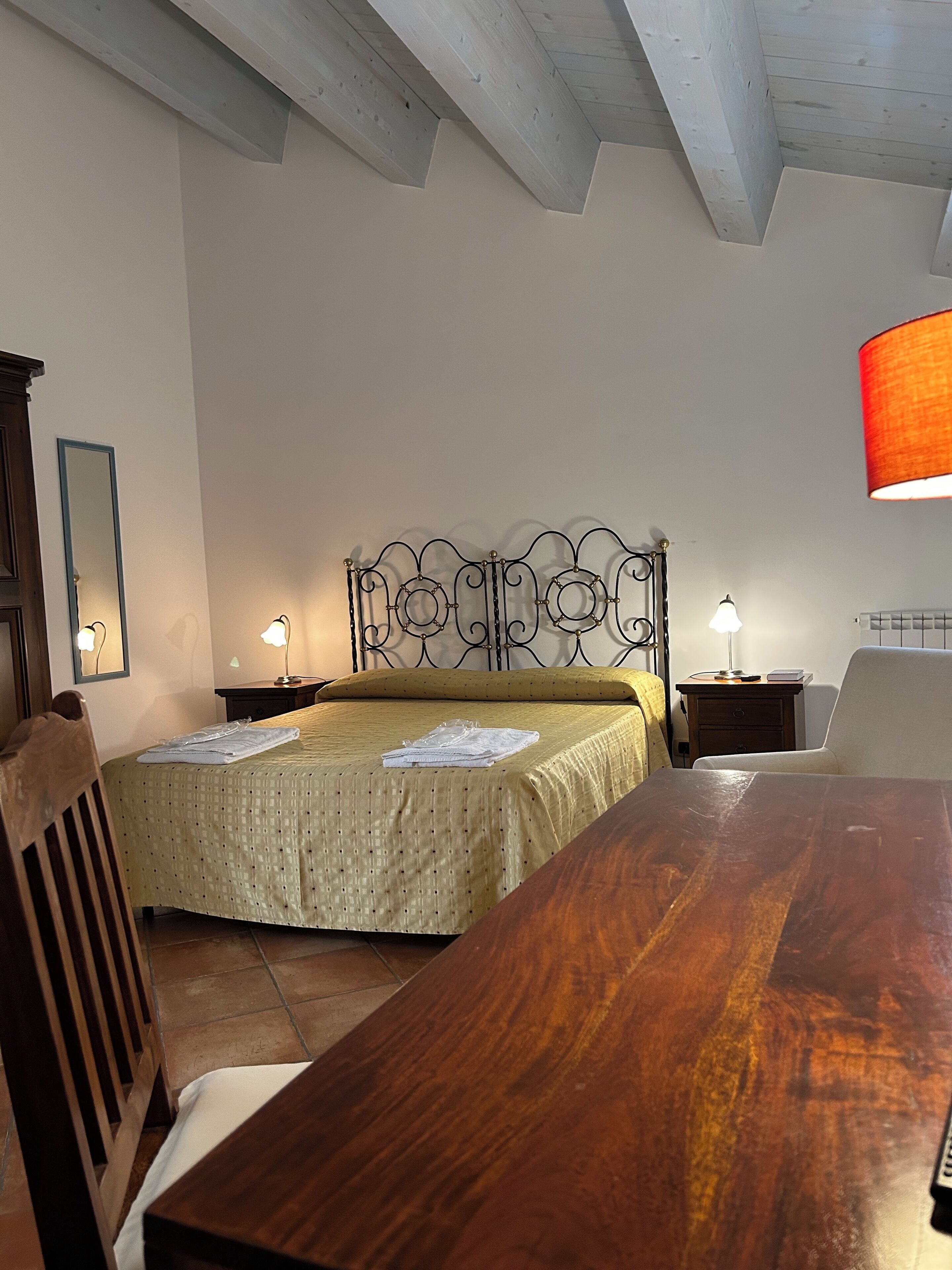 Un dormitorio con una cama de madera, una mesa de madera, una silla, una mesita de noche, una lámpara y un espejo.