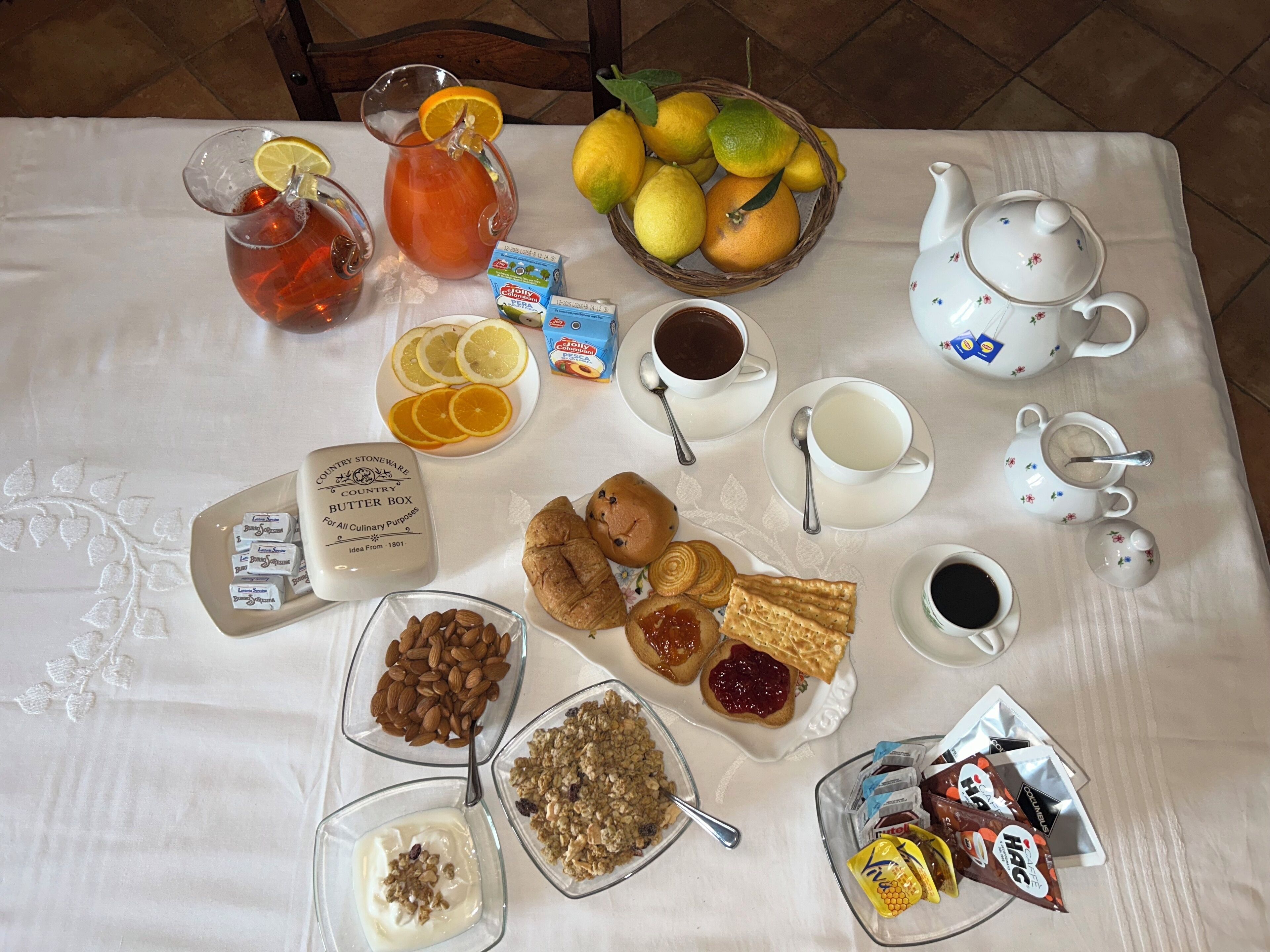 Una mesa de desayuno con diversos alimentos, una jarra de té, una tetera y una cesta de fruta.