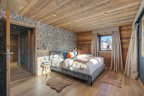 A log cabin-style bedroom with a bed, a small stool, a fur rug, and a window with curtains.