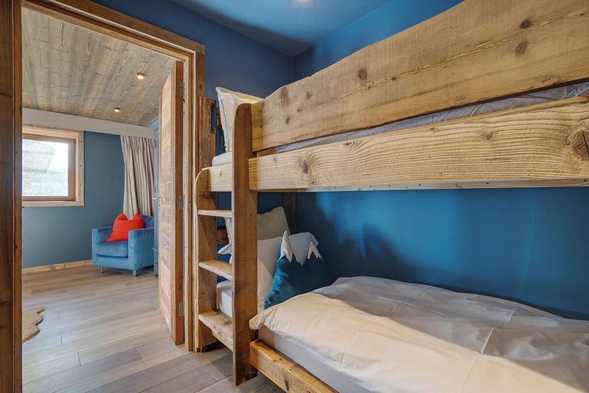 A bunk bed with a wooden frame, a blue wall, and a window with curtains.