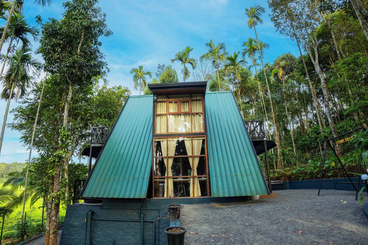 A building with a green roof and large windows, surrounded by tall trees and a clear sky.