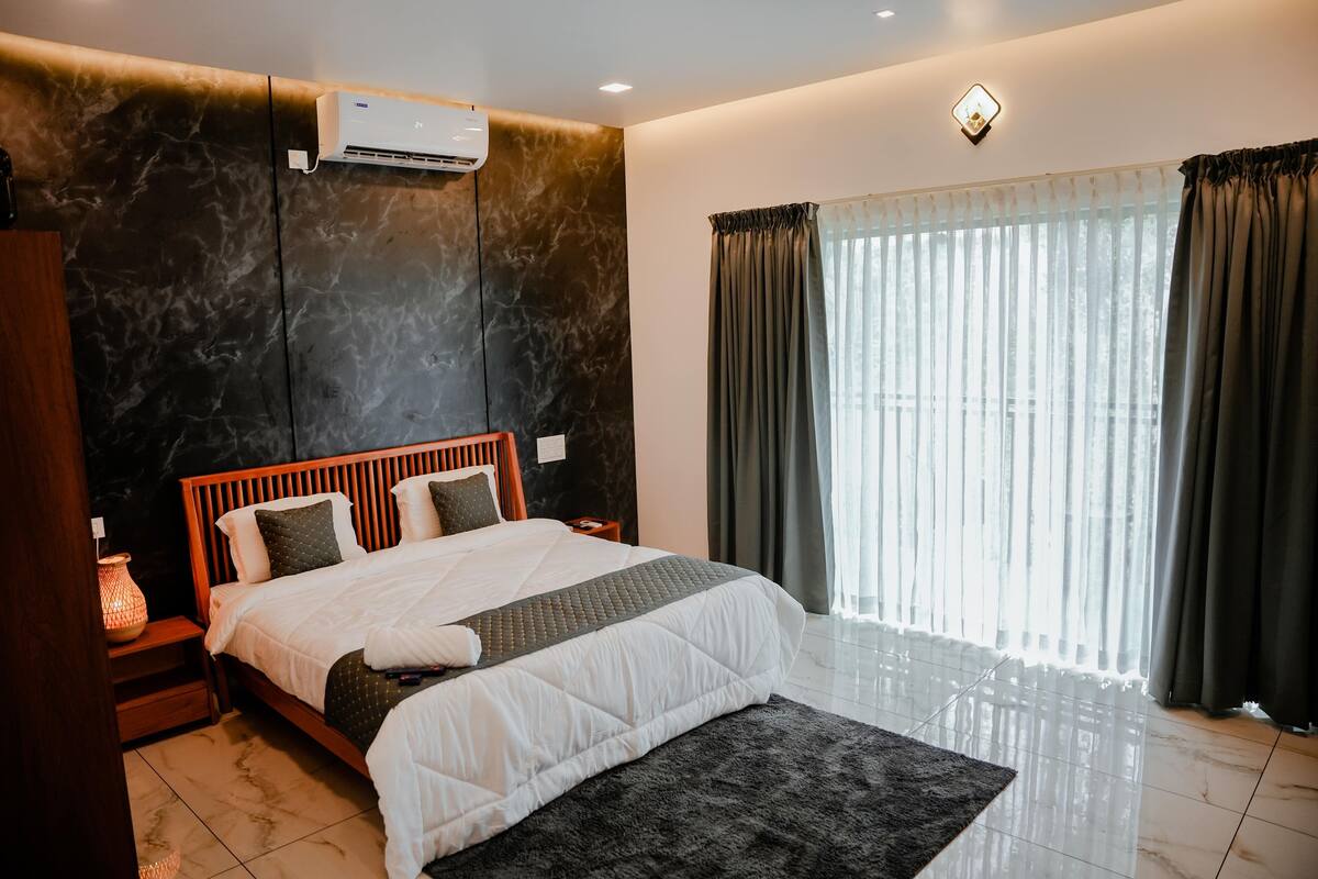 A modern bedroom with a large bed, a marble wall, an air conditioner, a nightstand with a lamp, and a window with curtains.
