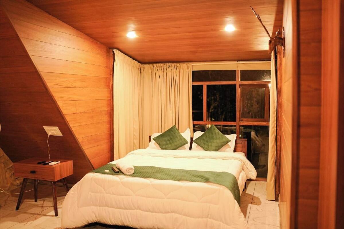 A bedroom with a wooden sloped ceiling, a bed with white bedding and green pillows, a bedside table, and a window with curtains.