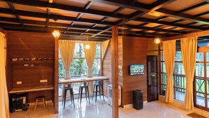 A room with a wooden ceiling, a dining table with chairs, a TV mounted on the wall, and large windows with curtains. - Ethnic Villa Wayanad (Sulthan Bathery)