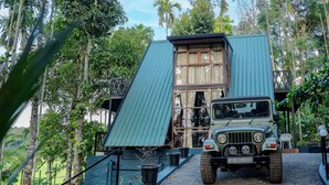 A green off-road vehicle parked in front of a building with a sloped roof, surrounded by tall trees. - Ethnic Villa Wayanad (Sulthan Bathery)
