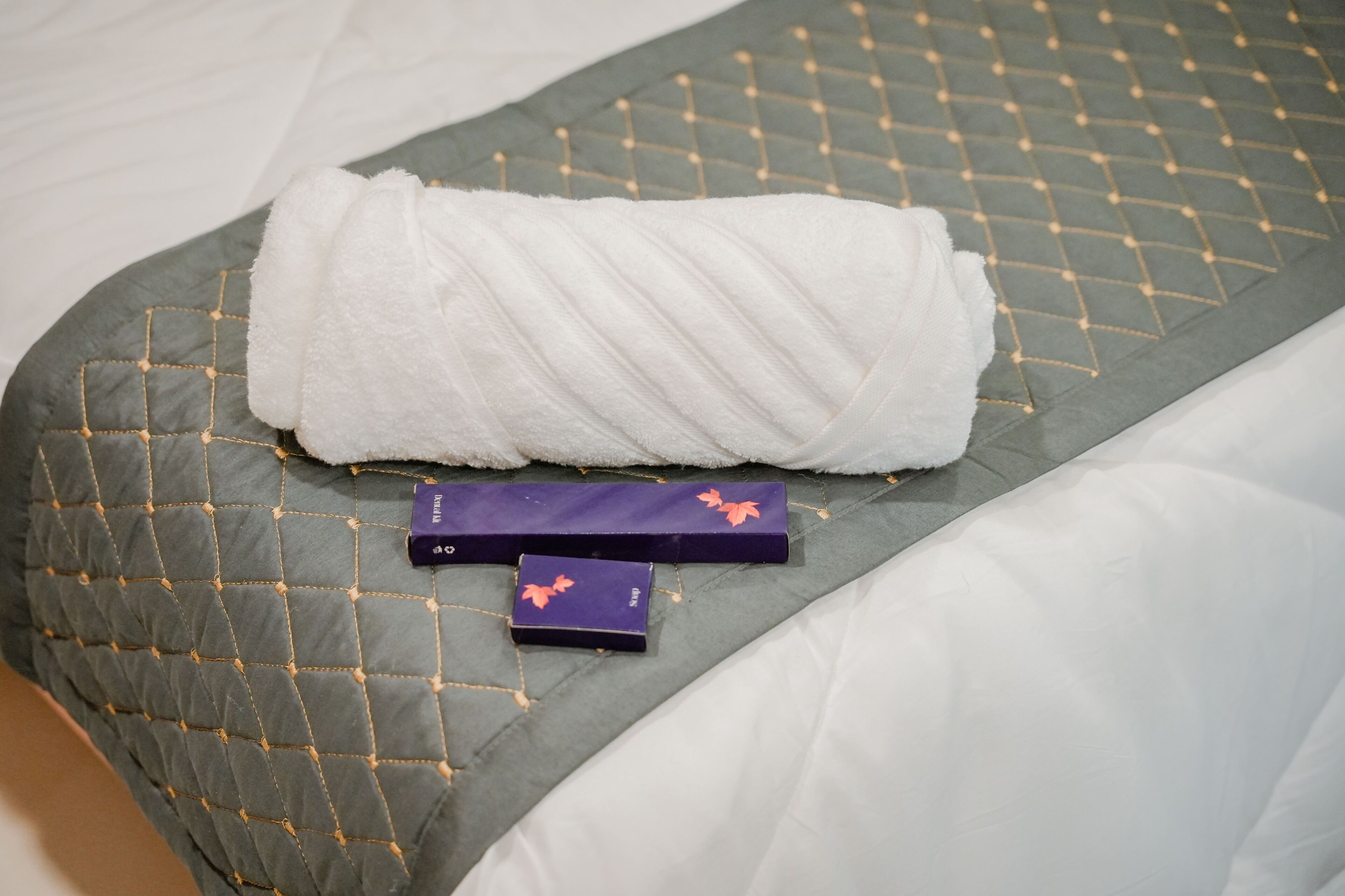 A neatly made bed with a quilted grey bedspread and a white towel placed on top.