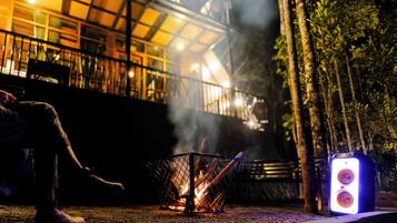 Ein Lagerfeuer mit Metallgrill, Lautsprecher und einer in der Nähe sitzenden Person.