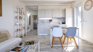 A modern, open-plan living and dining area with a sofa, coffee table, and chairs. A kitchen with white cabinets and a clock is visible in the background.