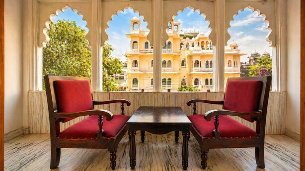 A room with a view of a building through arched windows, two wooden chairs with red cushions, and a marble floor. - The Crescent Udaipur | Boutique Hotel Near Lake Pichola (Udaipur)