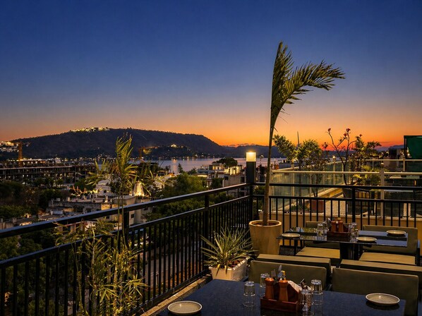 Une terrasse sur le toit, avec des tables et des chaises, d’où l’on peut admirer un panorama sur la ville et une colline au coucher du soleil.