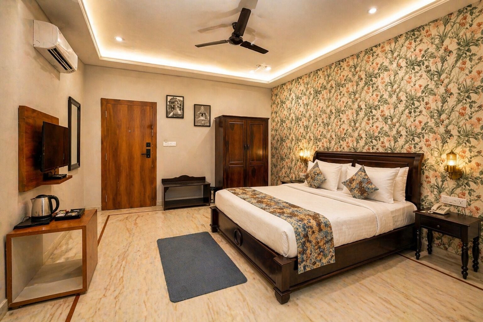 A bedroom with a bed, a ceiling fan, a TV, a wardrobe, a desk with a kettle, and a floral wallpaper.