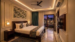 A hotel room with a large bed, a ceiling fan, a TV, and a view of the cityscape at dusk. - The Crescent Udaipur | Boutique Hotel Near Lake Pichola (Udaipur)