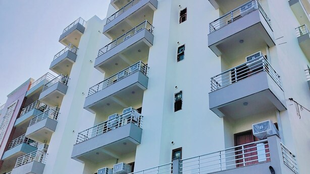 A multi-story residential building with balconies, air conditioning units, and a storefront at the ground level. - Xact Homes  (Noida)