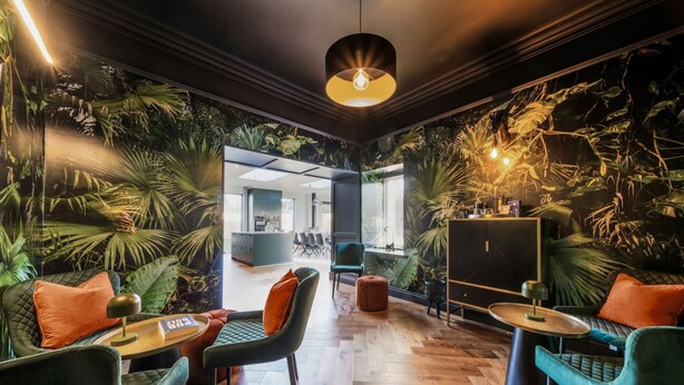 A modern interior with a tropical theme, featuring a dining area with chairs and tables, a kitchen in the background, and a large plant wall.