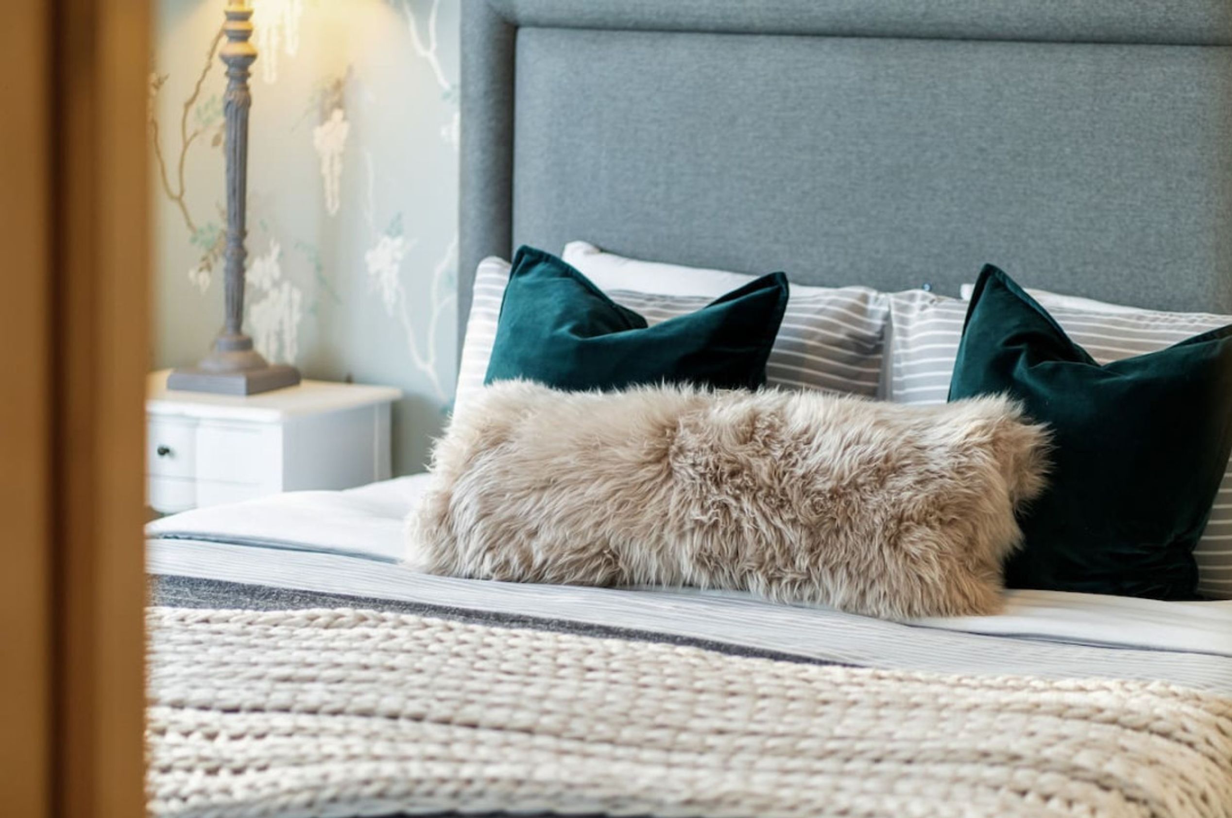 A neatly made bed with a fluffy pillow and two dark pillows, a bedside lamp, and a wall with a floral pattern.