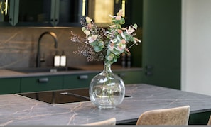 Um vaso de vidro transparente com um arranjo floral sobre uma bancada de mármore em uma cozinha moderna com armários verdes e uma pia de aço inoxidável.