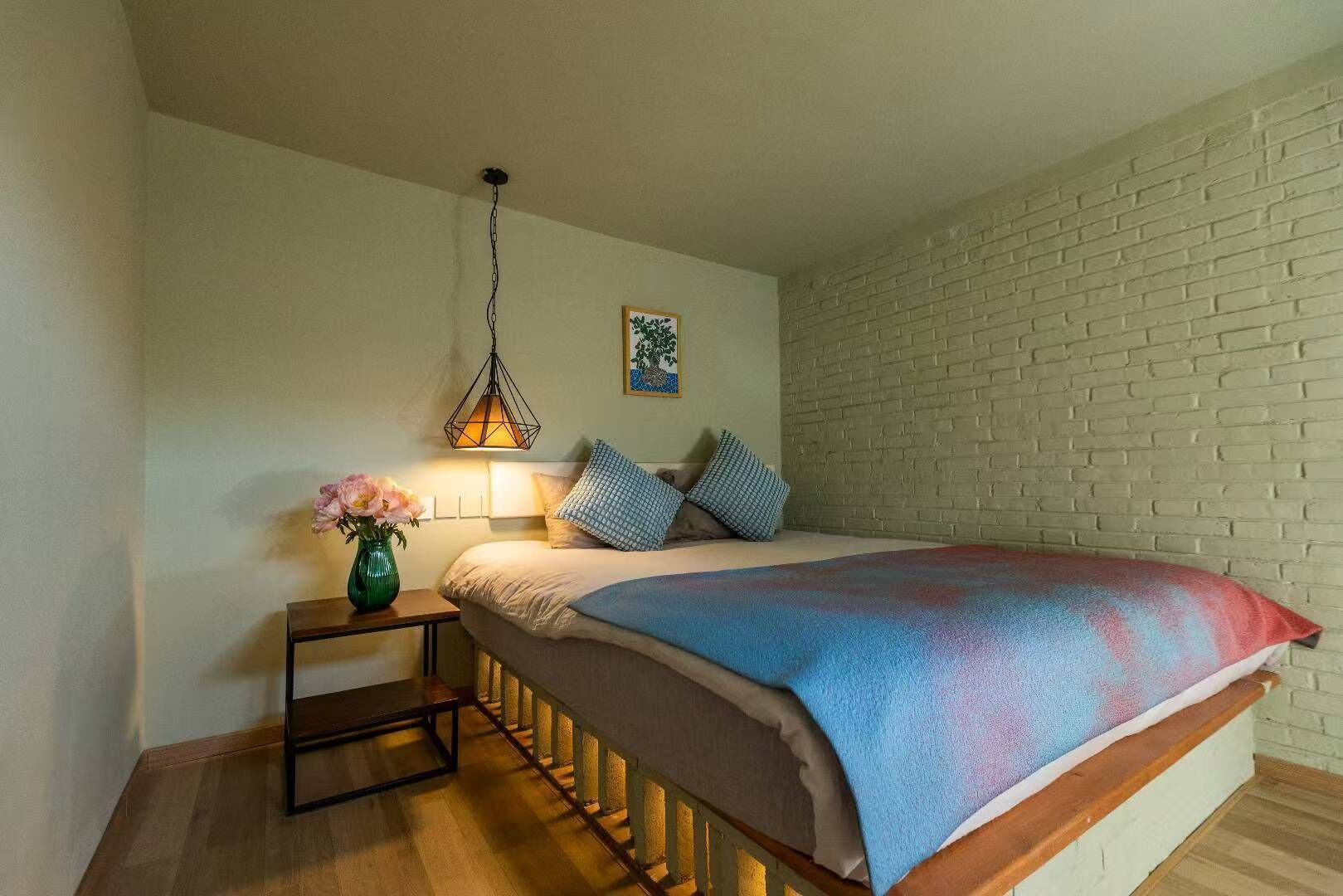 A bedroom with a bed, a nightstand, a hanging lamp, a wall-mounted picture, and a brick wall.