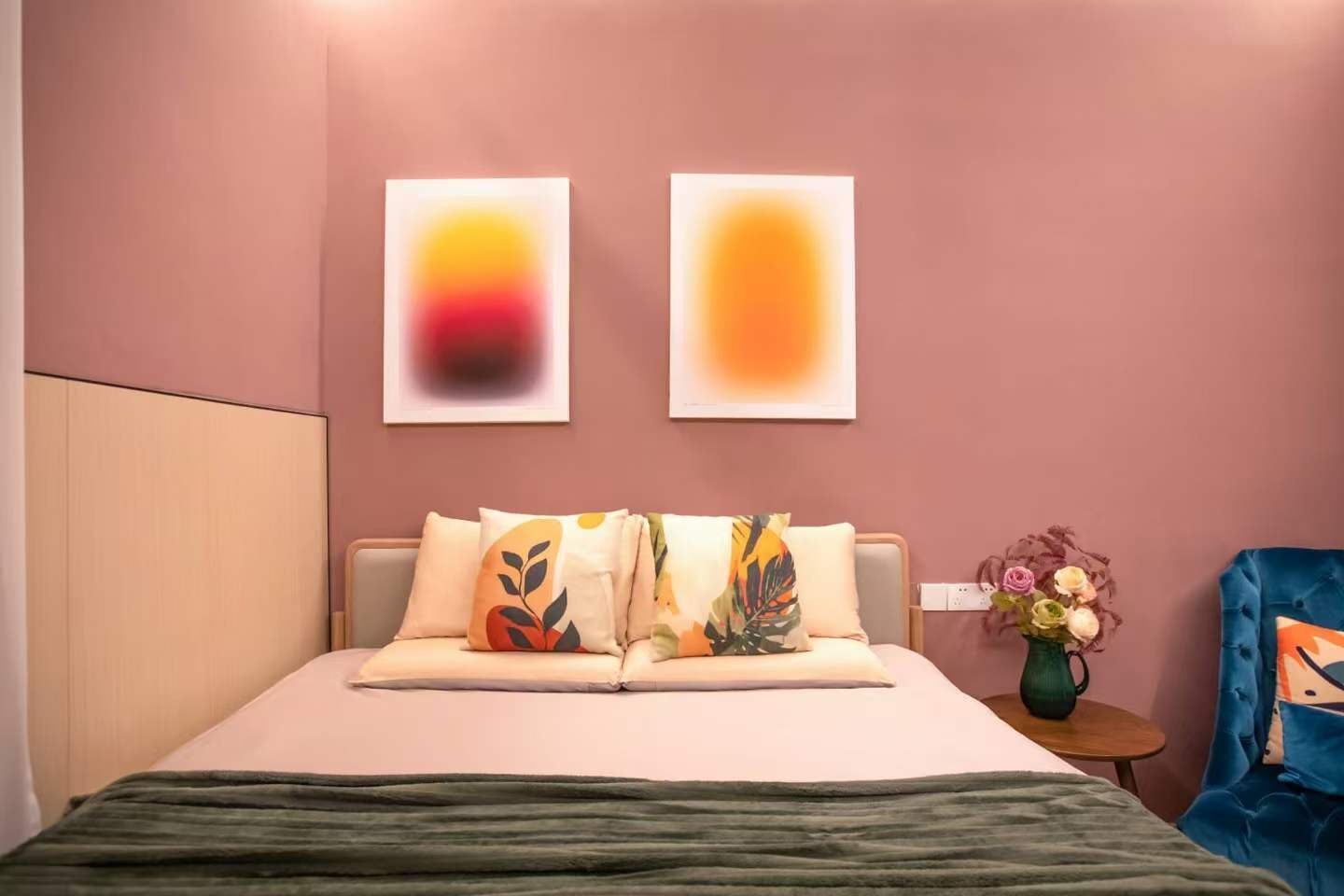 Un dormitorio con cama, almohadas, una mesita de noche, un florero con flores y dos cuadros abstractos en la pared.