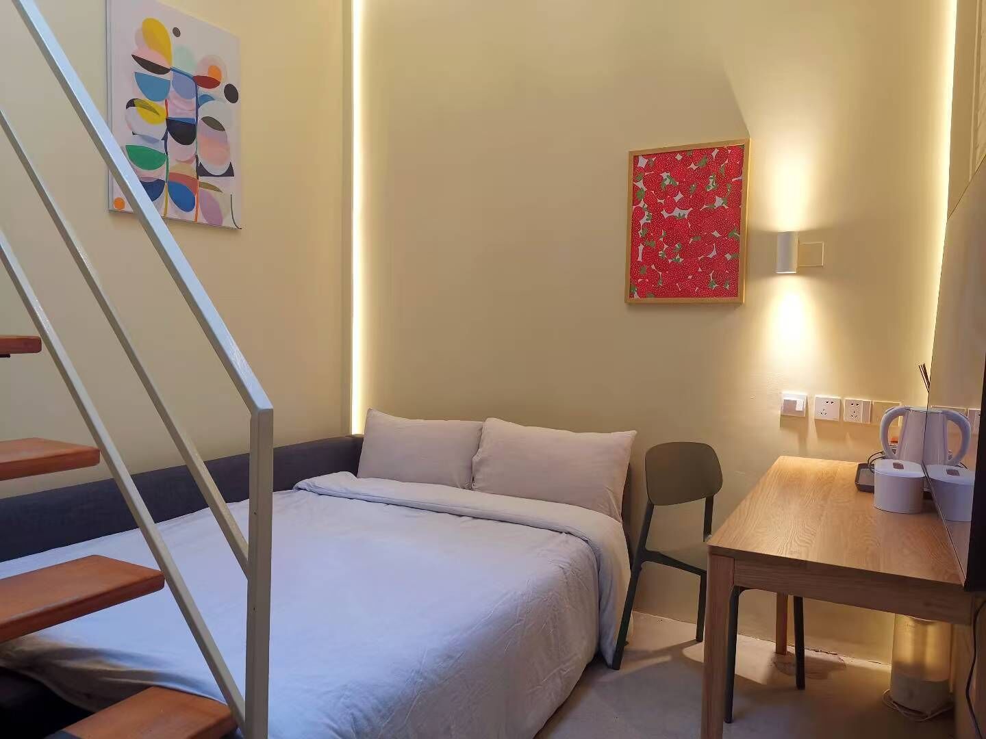 Un dormitorio con cama, escritorio, silla, escalera y dos obras de arte en la pared.