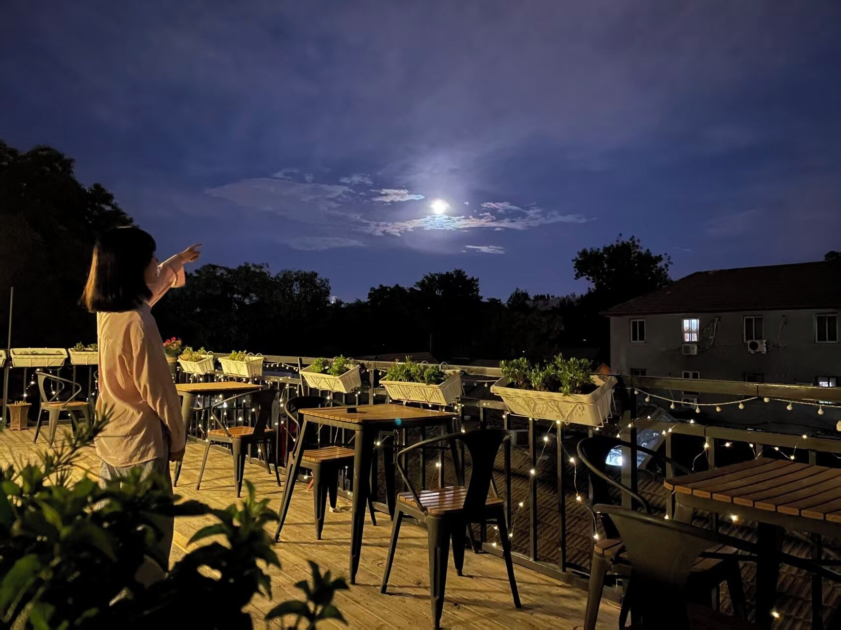 Un área de comedor al aire libre con mesas y sillas, rodeada de plantas en macetas, bajo un cielo nocturno con una luna visible.