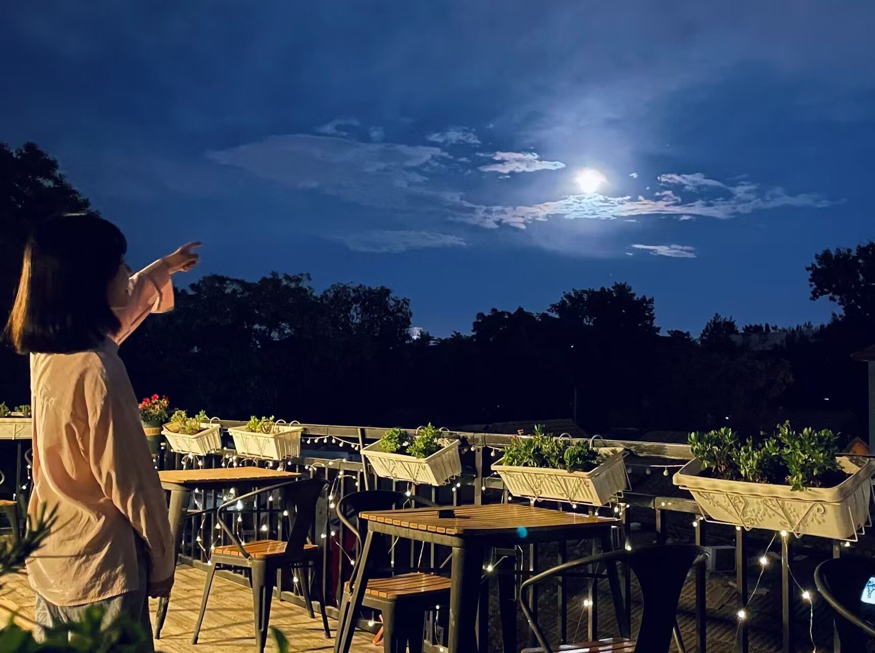 An outdoor seating area with tables and chairs, surrounded by potted plants, under a night sky with a visible moon.