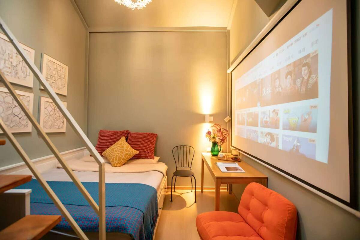 Une chambre d’hôtel compacte comprenant un lit, un bureau avec une chaise, une petite table avec un vase et un écran de projection affichant des images.