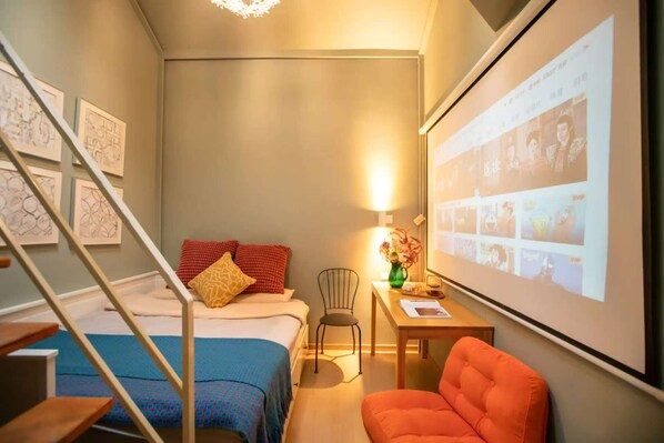 A compact hotel room with a bed, a desk with a chair, a small table with a vase, and a projector screen displaying images. - Beijing Drum Tower Loft Courtyard Hotel (Beijing)