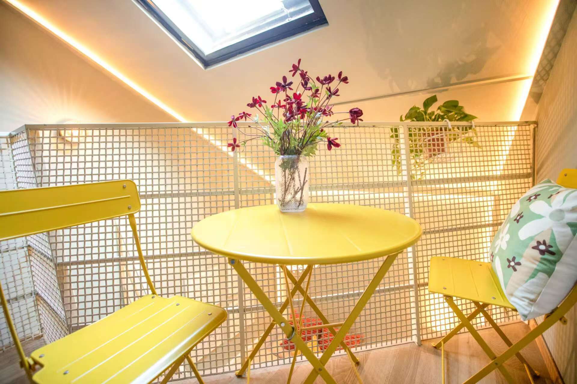 Una mesa amarilla con un florero, dos sillas amarillas y una partición de malla en una habitación con techo inclinado y tragaluz.
