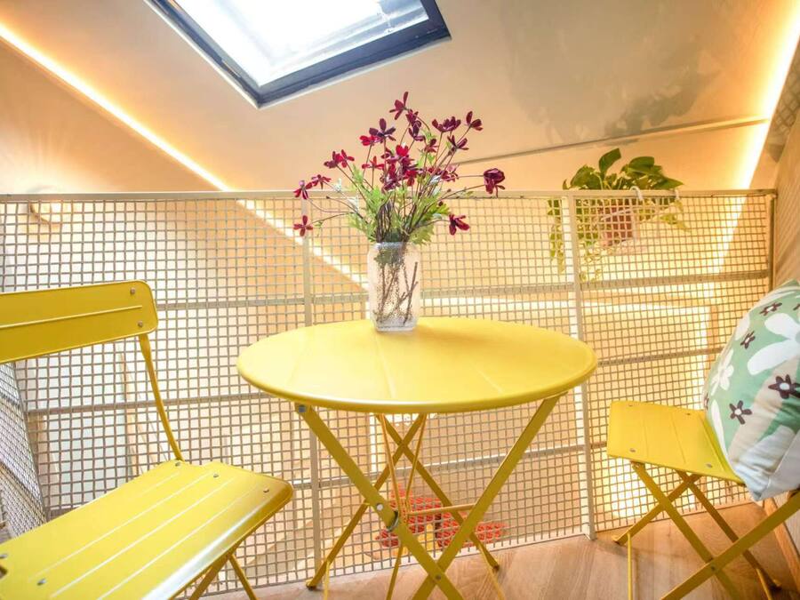 Una mesa amarilla con un florero, dos sillas amarillas y una partición de malla en una habitación con techo inclinado y tragaluz.