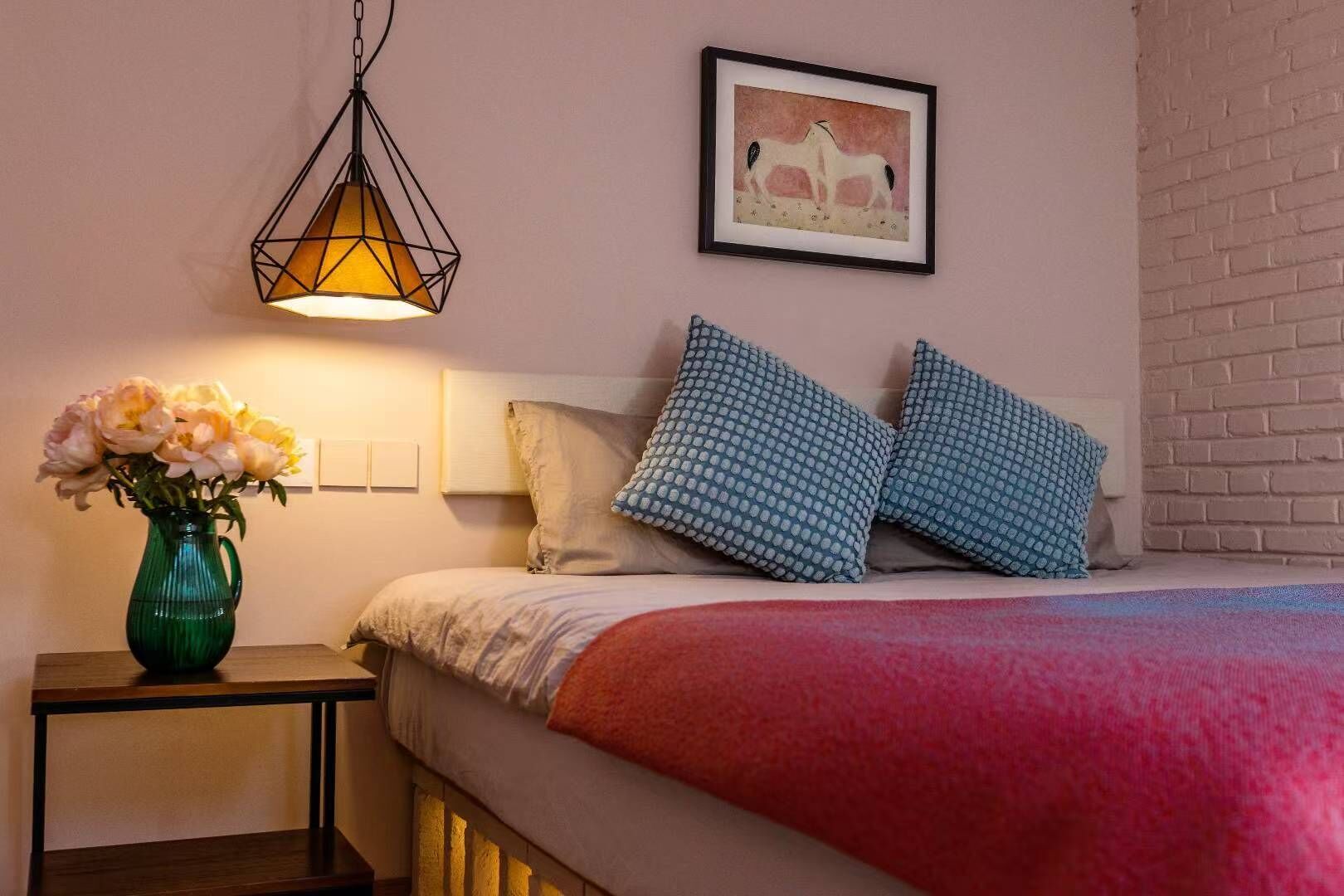 A bedroom with a bed, pillows, a vase with flowers, a hanging light fixture, and a framed picture on the wall.