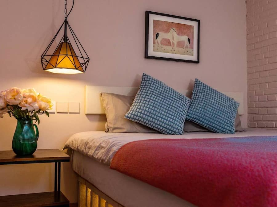 A bedroom with a bed, pillows, a vase with flowers, a hanging light fixture, and a framed picture on the wall.