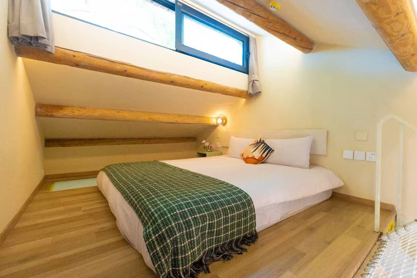 Une chambre avec un plafond en pente, des poutres en bois, un lit recouvert d’une couverture à carreaux verts, une table de chevet et une fenêtre avec des rideaux.