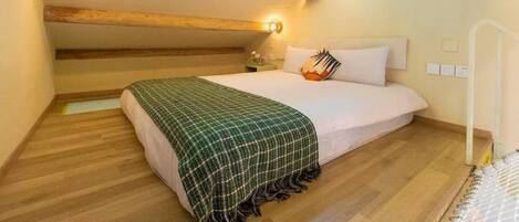 Une chambre avec un plafond en pente, des poutres en bois, un lit recouvert d’une couverture à carreaux verts, une table de chevet et une fenêtre avec des rideaux.